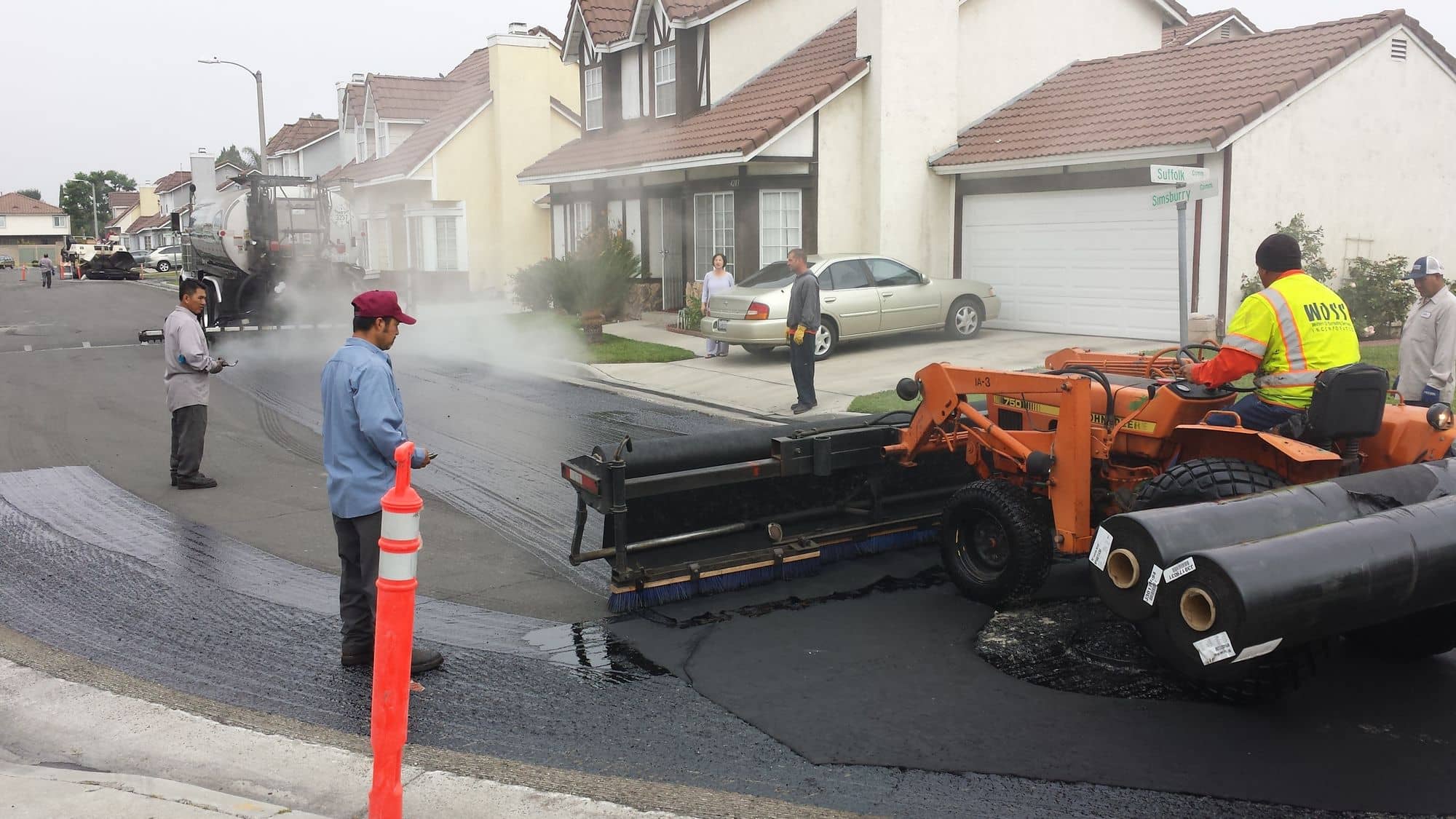 Crew member spraying the street surface while a tractor-mounted rig unrolls black paving fabric along a residential curb